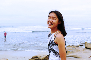 Young Asian woman smiles at the camera while walking on rock near the beach on a cloudy day.