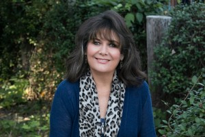 Woman in a dark blue sweater with a leopard print scarf smiling at the camera with a natural background behind her.