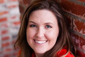 A smiling woman, brown hair, in front of a brick wall. Kristin Pedroza