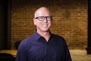 White man wearing glasses and a blue shirt smiling at the camera in front of a brick wall background