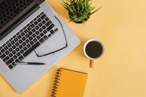 Laptop open on a yellow desktop with reading glasses, coffee cup, a notebook and a plant