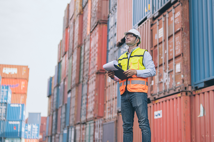 Worker in vest holding safety log among shipping containers
