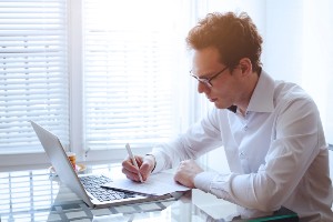 Man with curly brown hair sitting at a desk writing with a laptop open