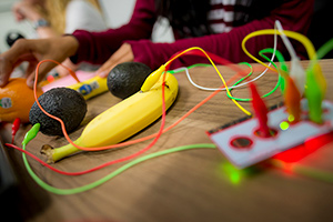 Wires plugged into both a banana and a couple of avocados, with a young woman in the background