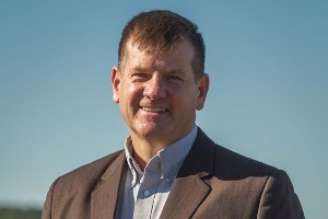 Michael McGinnis, middle age, white man wearing a light blue button down shirt and a brown jack, smiling